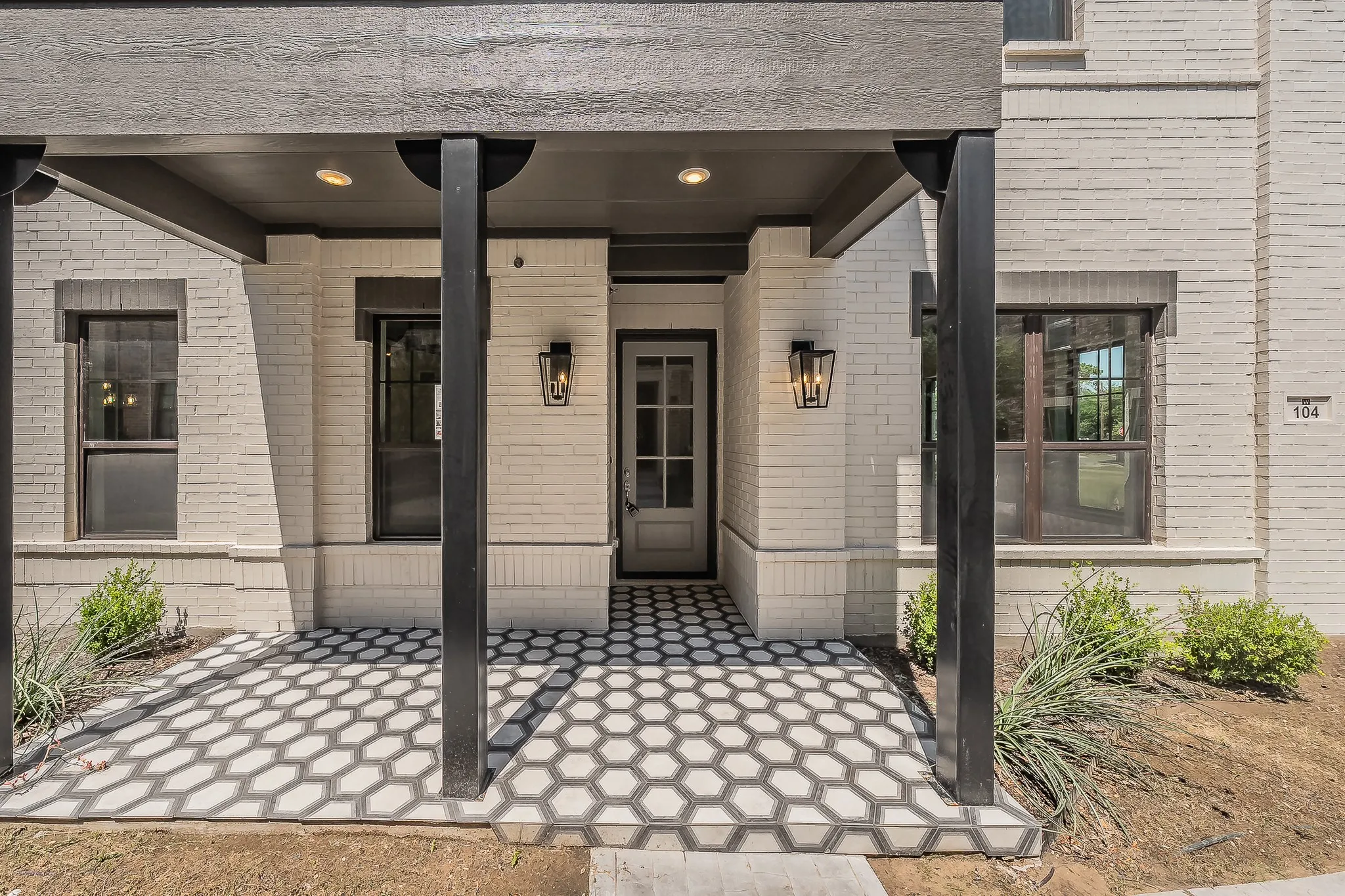 Entryway featuring a covered porch, white brick exterior, black hexagonal tile flooring, and two wall-mounted lanterns flanking the front door