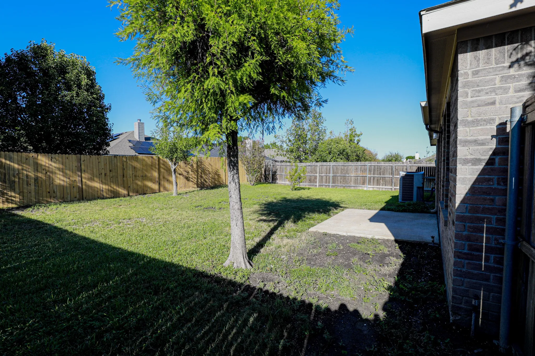 Expansive grass area with a privacy fence, mature trees, a concrete patio, and a brick exterior