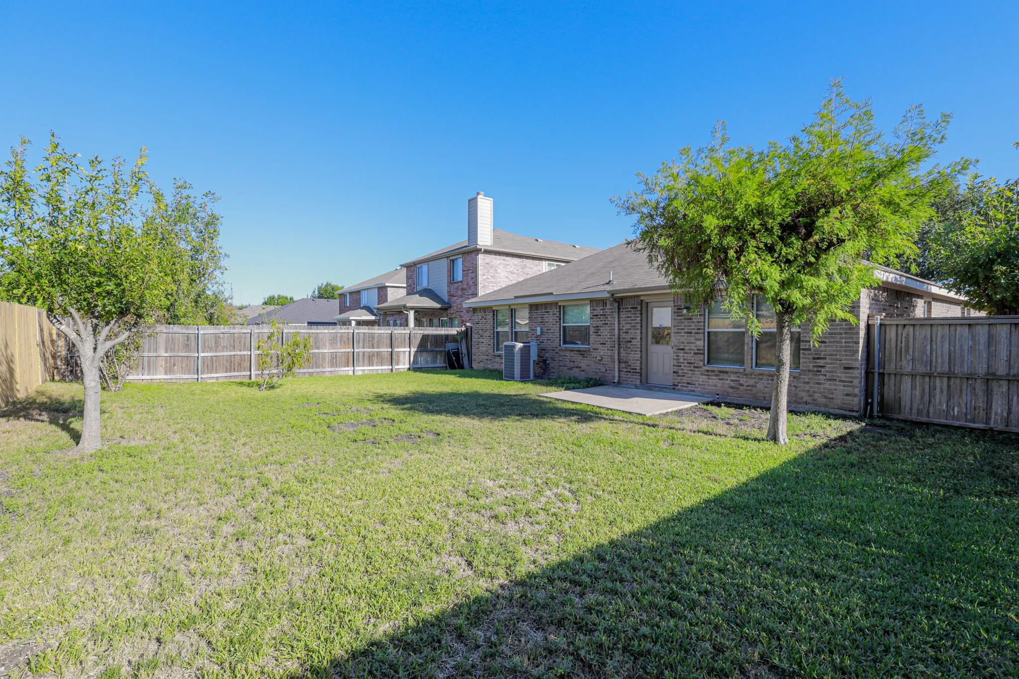 Fenced backyard with a concrete patio, mature trees, and a brick exterior