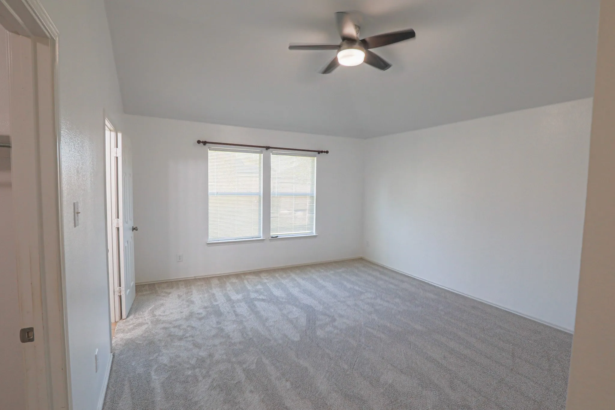Spacious room featuring vaulted ceiling, ceiling fan with integrated lighting, two windows with blinds, and light-toned carpet flooring
