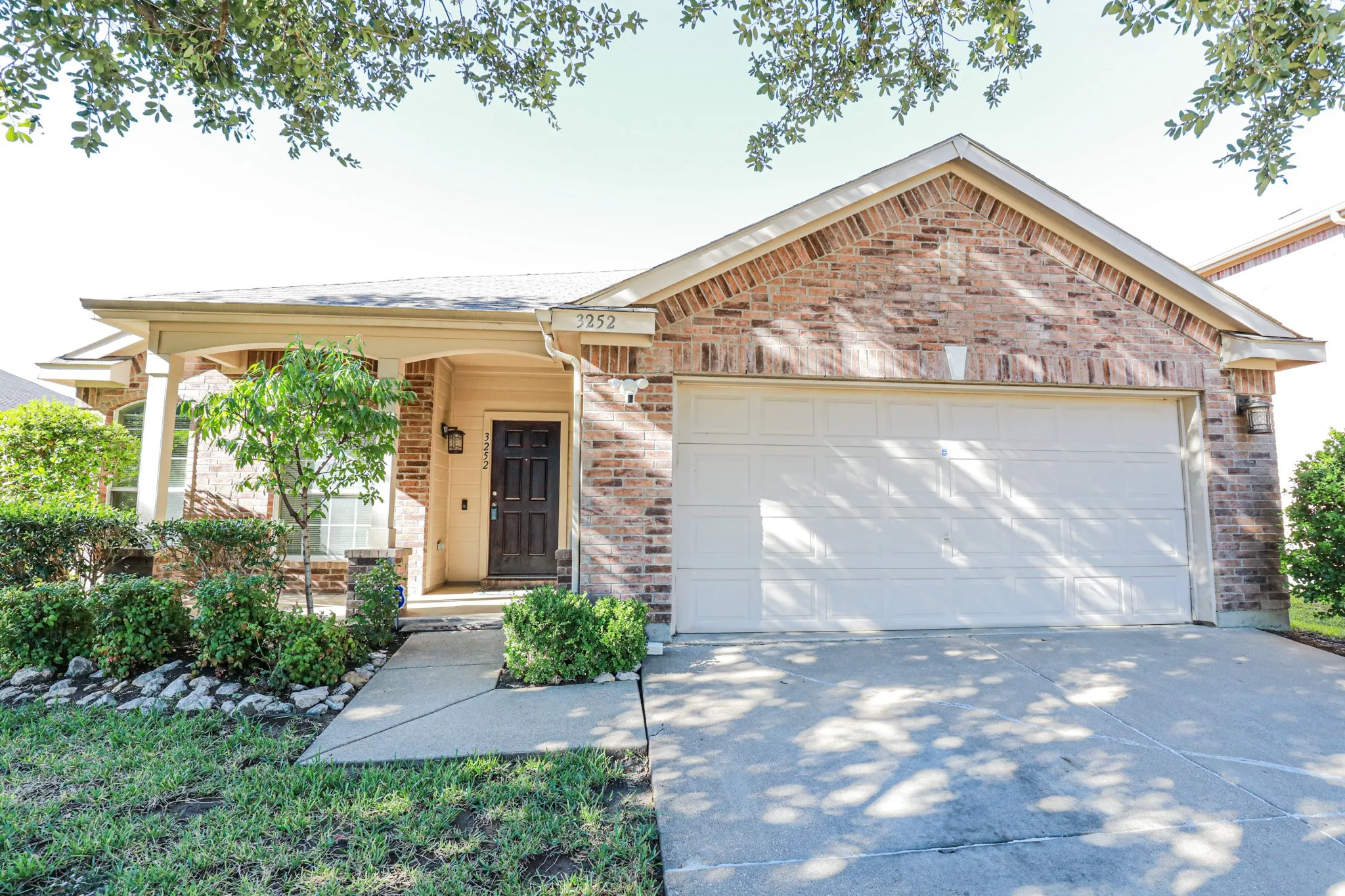 Brick exterior with a covered entry, attached garage, concrete driveway, and mature landscaping