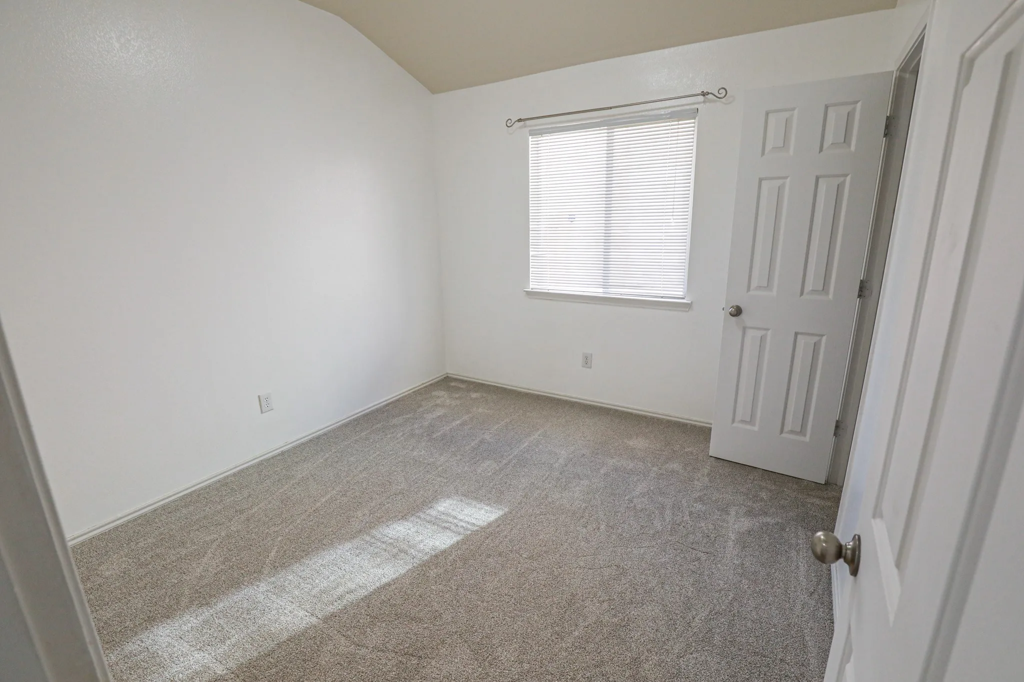 Neutral carpet flooring with white walls, a single window with blinds, and a white paneled door