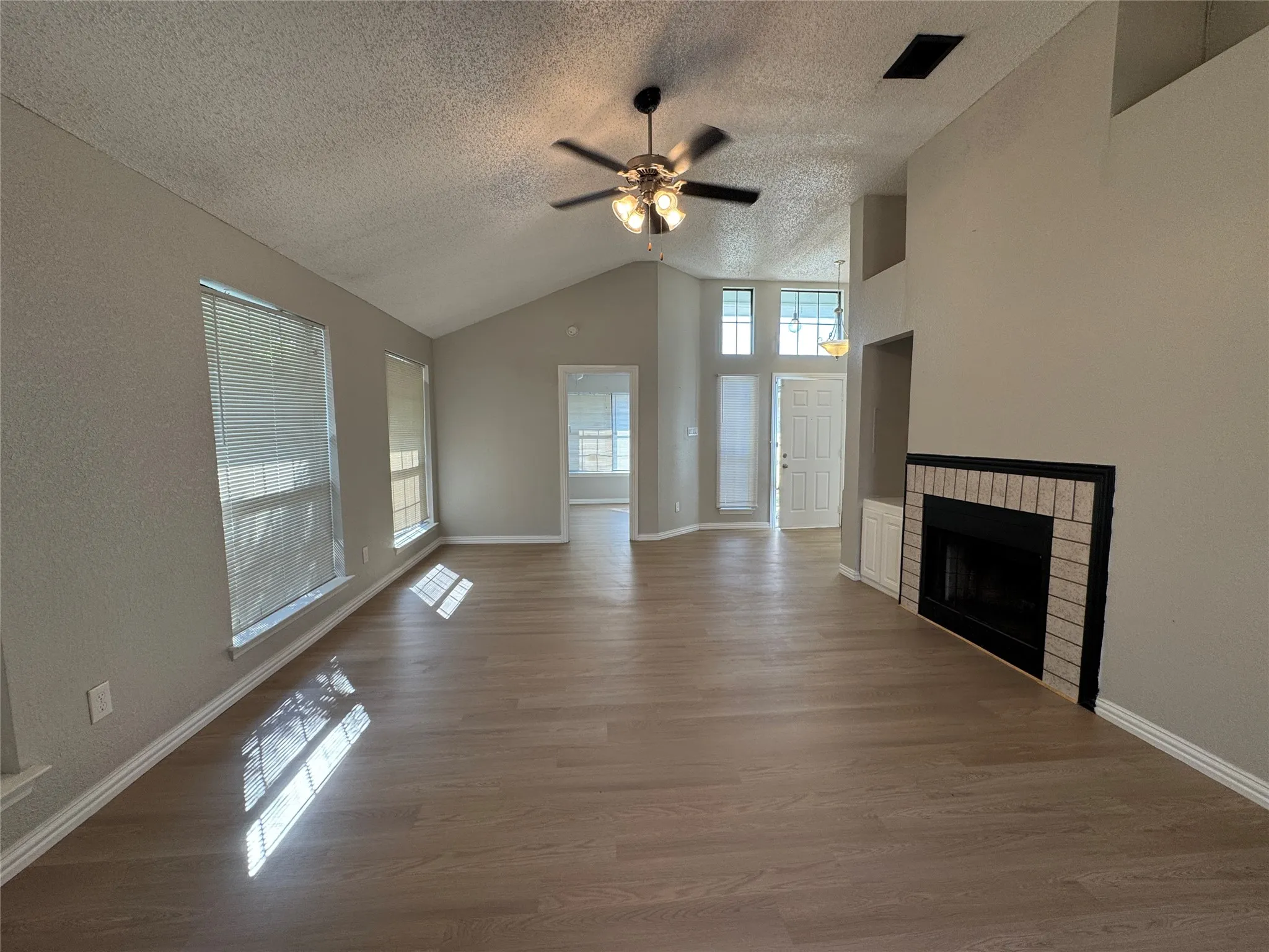 Vaulted ceiling with a ceiling fan, wood-finish flooring, multiple windows, and a fireplace with a light-colored brick surround