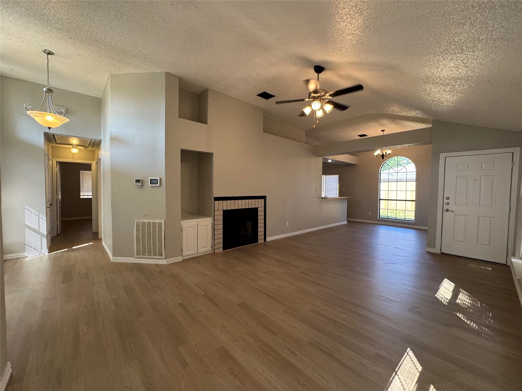 Expansive open-concept living area featuring wood-finish flooring, a fireplace with a brick surround, built-in shelving, vaulted ceilings, and an arched window