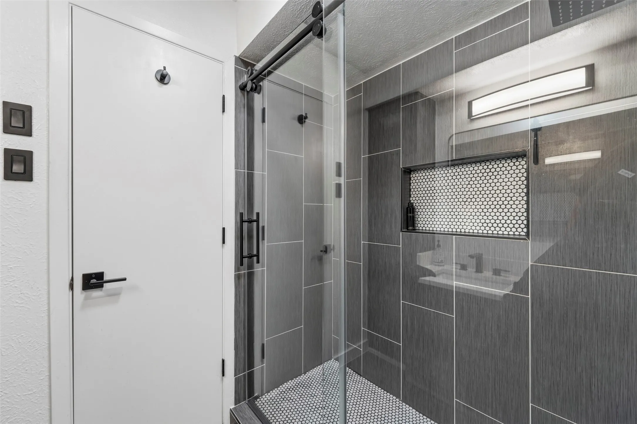 Shower with dark gray rectangular tile, black penny tile flooring, built-in recessed shelf with penny tile, and sliding glass enclosure with black hardware