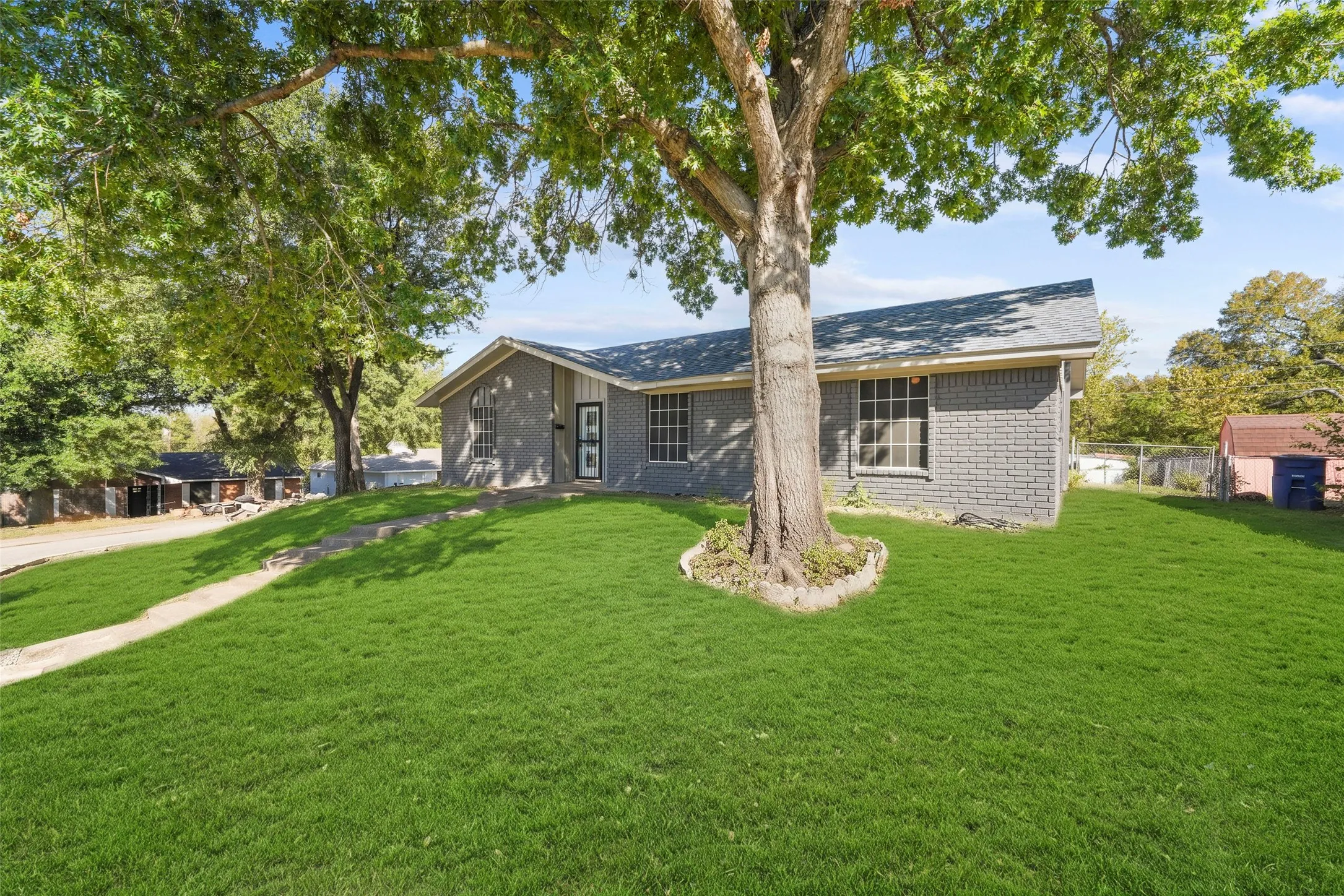 Gray brick exterior, composition shingle roof, mature front yard tree, concrete walkway