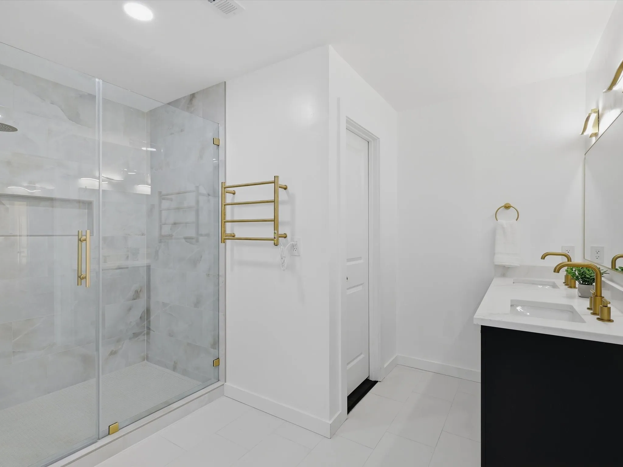 Spacious bathroom featuring a glass-enclosed shower with stone-finish tile, a dual vanity with a white countertop and gold-finish fixtures, and gold-finish lighting