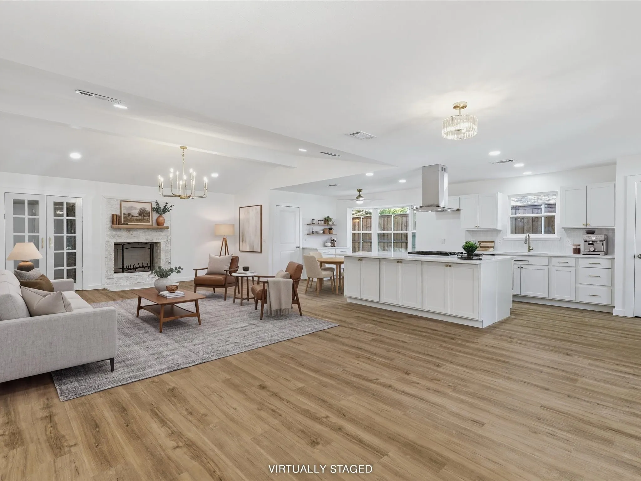 Open-concept living space featuring wood-finish flooring, a white kitchen island with counter seating, a stainless steel range hood, a fireplace with a white brick surround, and white kitchen cabinetry