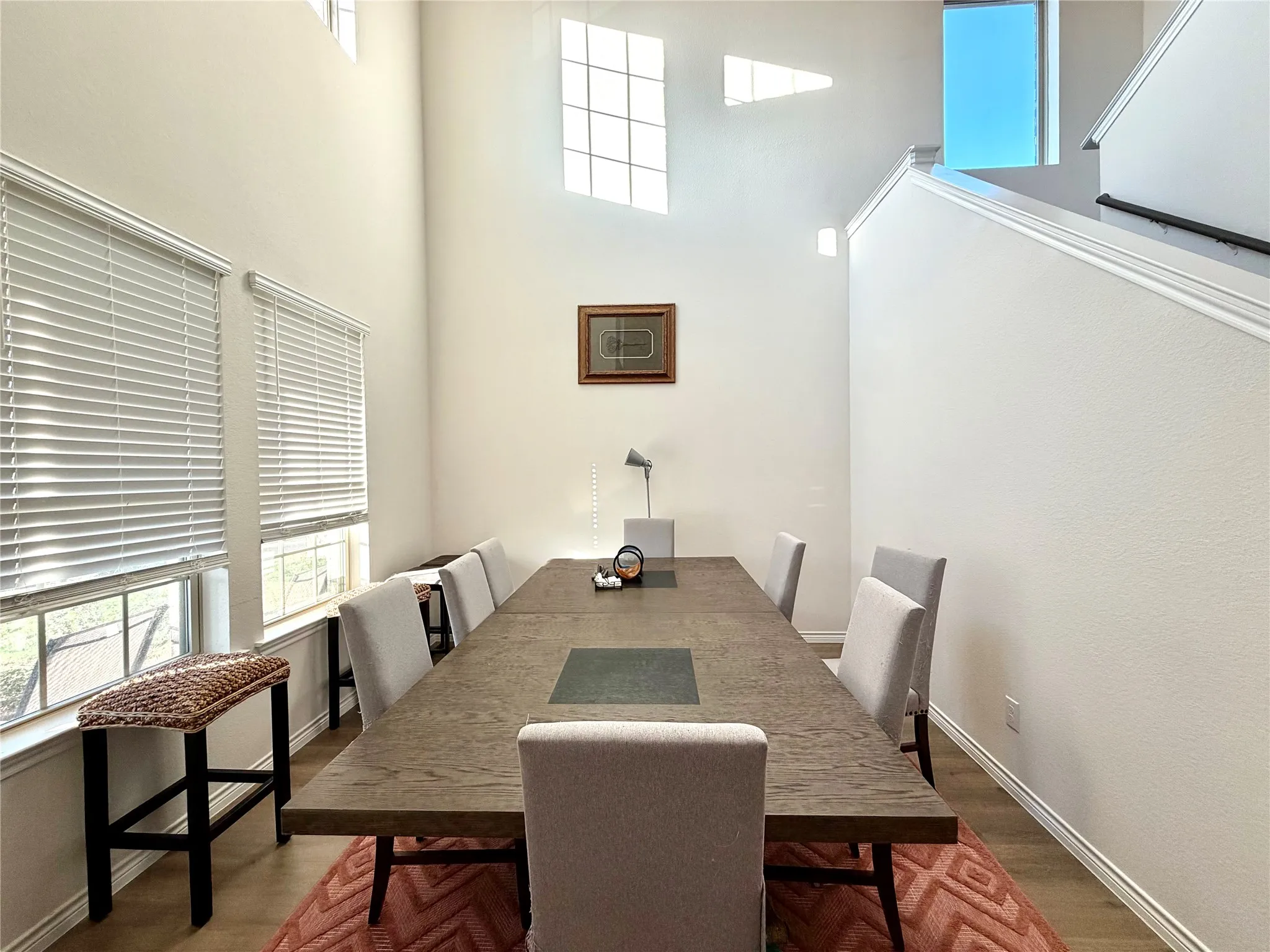 Vaulted ceiling with high transom windows and upper-level windows, wood-finish flooring, two large windows with blinds, and a staircase with a handrail