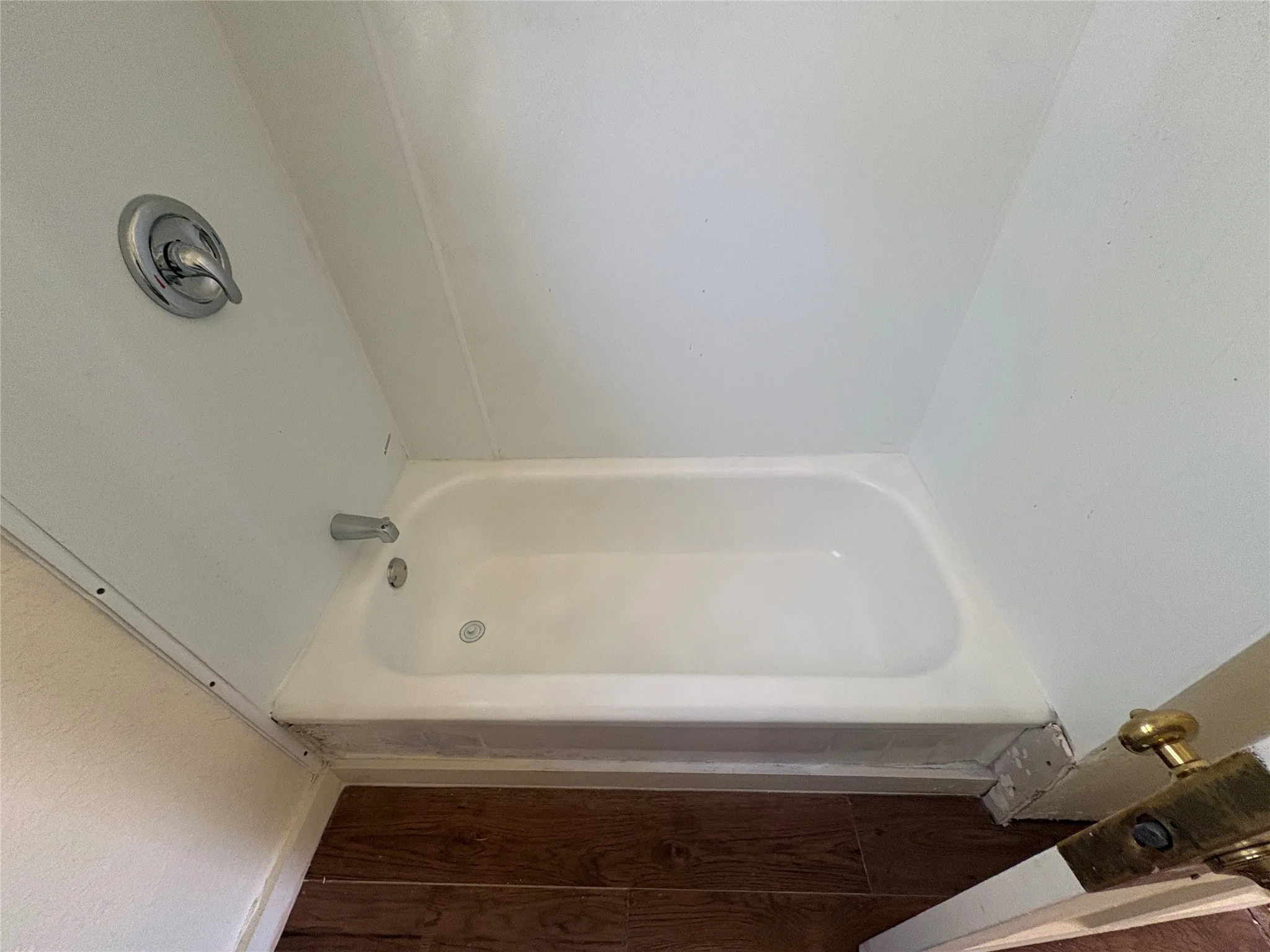 White bathtub and shower surround with polished chrome fixtures and wood-finish flooring