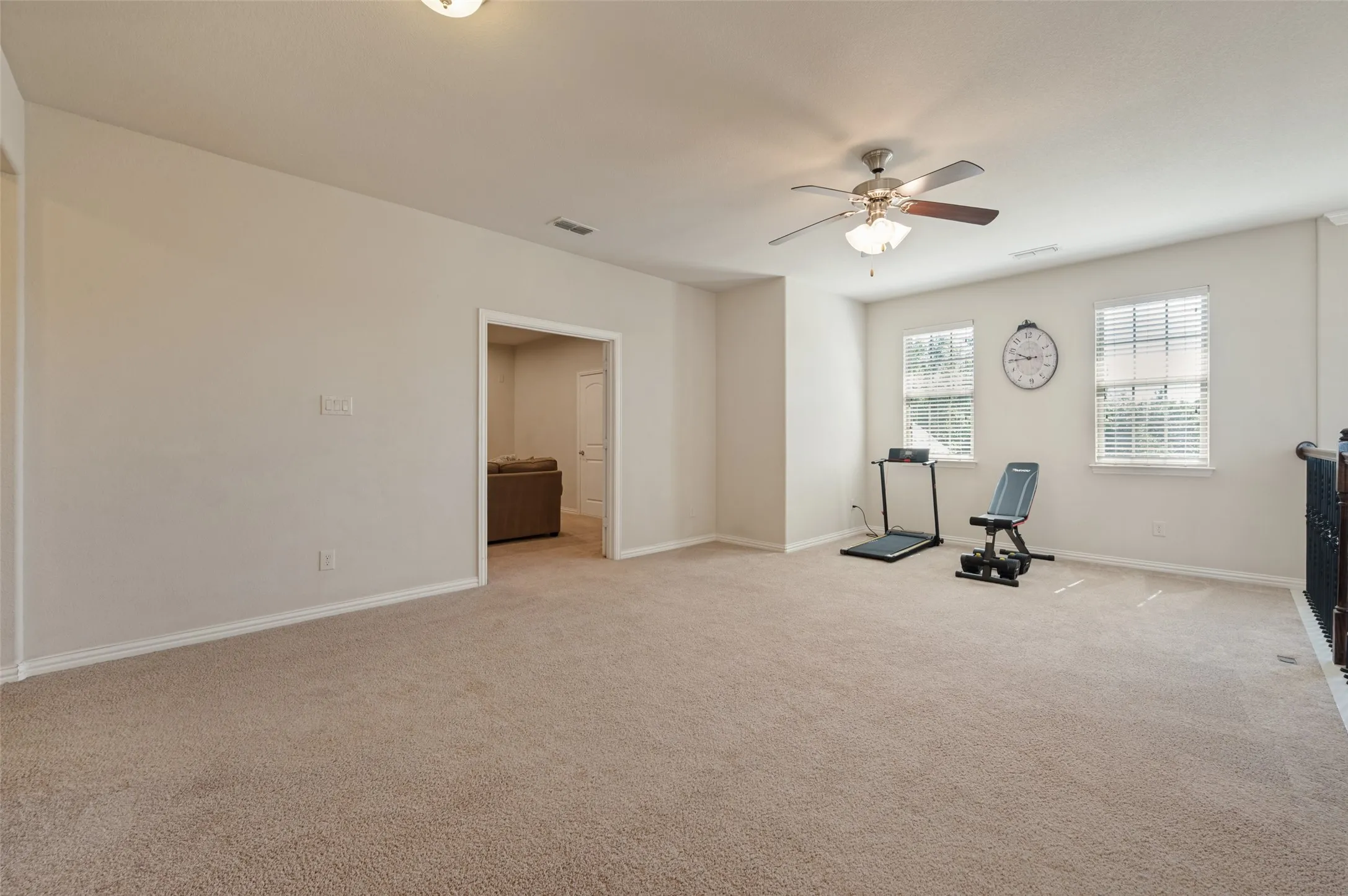 Spacious carpeted room featuring a ceiling fan with integrated lighting, two windows with blinds, and an open doorway leading to another room