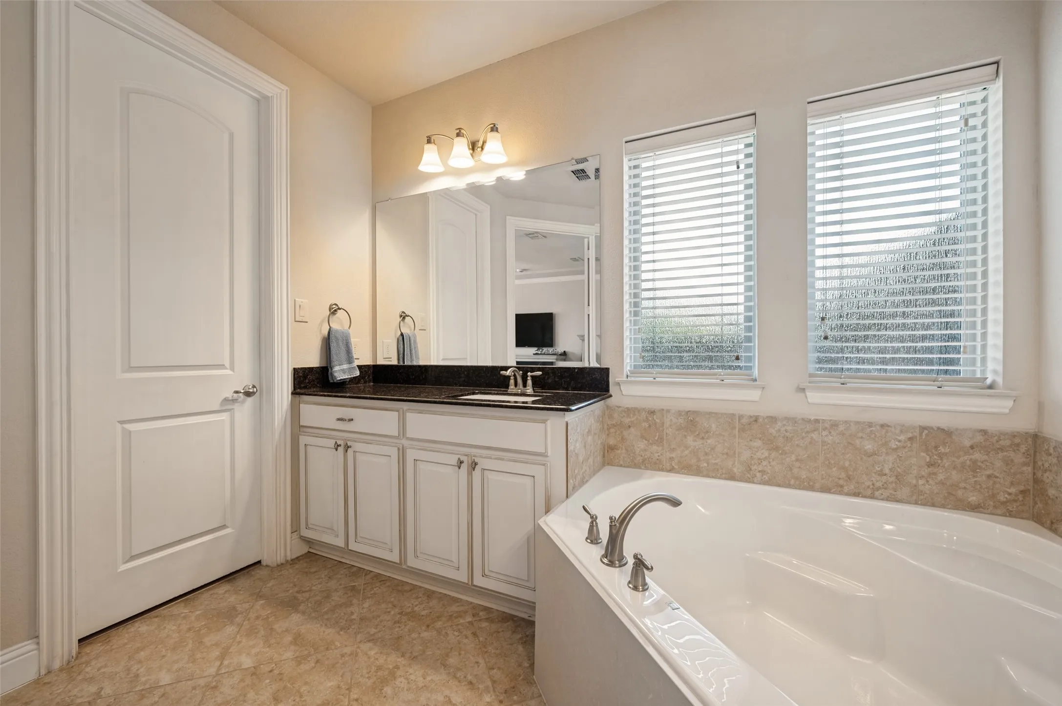 Soaking tub with brushed nickel fixtures, dual windows with blinds, single vanity with dark countertop, light-colored cabinetry, and tile flooring