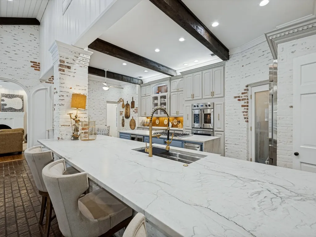 Expansive kitchen island with a stone-finish countertop, white-washed brick walls, vaulted ceiling with exposed wood beams, built-in stainless steel double ovens, and light-colored cabinetry
