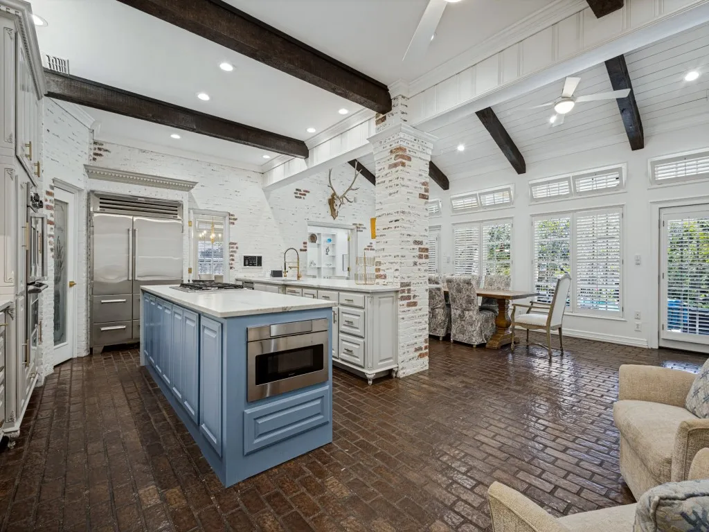 Spacious kitchen featuring exposed wood beams, brick accent walls, a kitchen island with built-in microwave, stainless steel appliances, and brick flooring