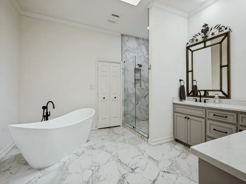 Freestanding soaking tub with a matte black faucet, glass-enclosed shower with tile surround, double vanity with light gray cabinetry, light-toned countertops, and marble-look tile flooring
