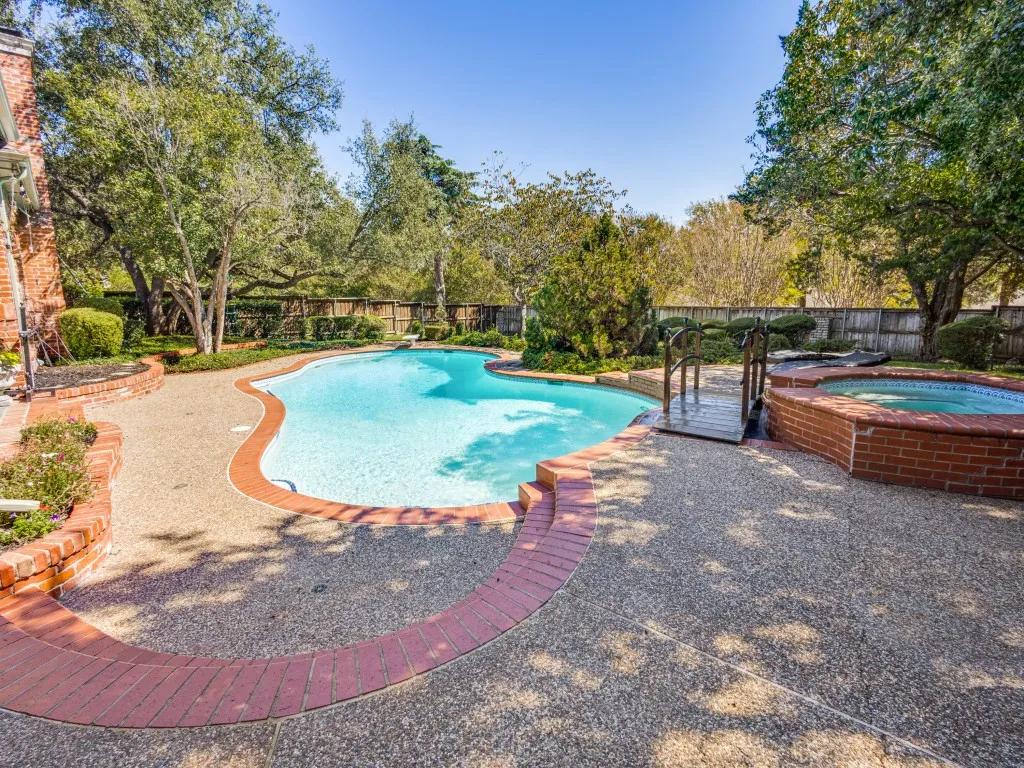 Curved swimming pool featuring an integrated spa, exposed aggregate decking, brick coping, and mature landscaping