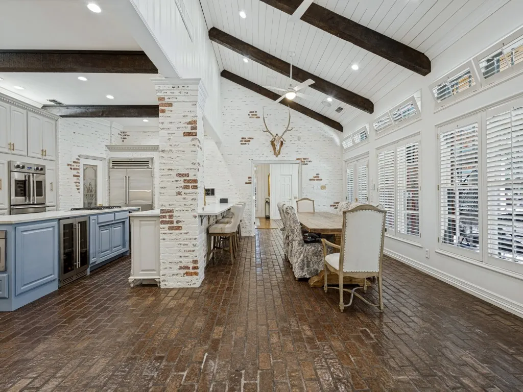 Vaulted ceiling with exposed wood beams, brick-finish flooring, white-painted brick walls, and extensive window shutters