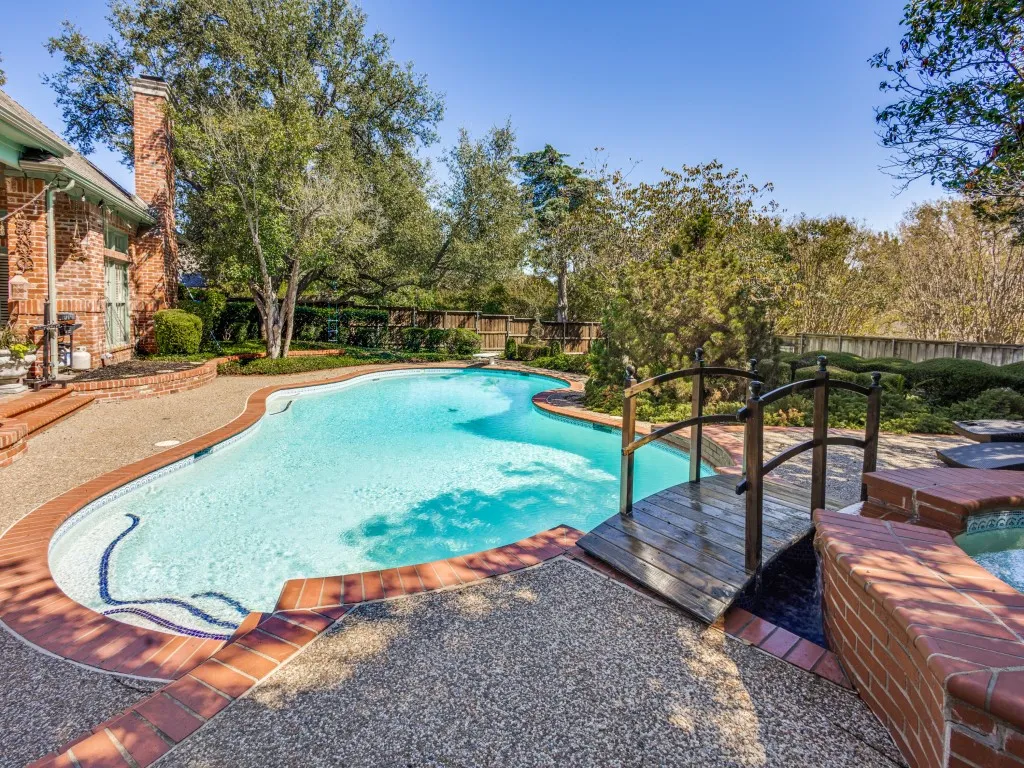 Curved swimming pool with mosaic tile details, a decorative wooden bridge, brick-lined spa, aggregate concrete decking, and mature landscaping