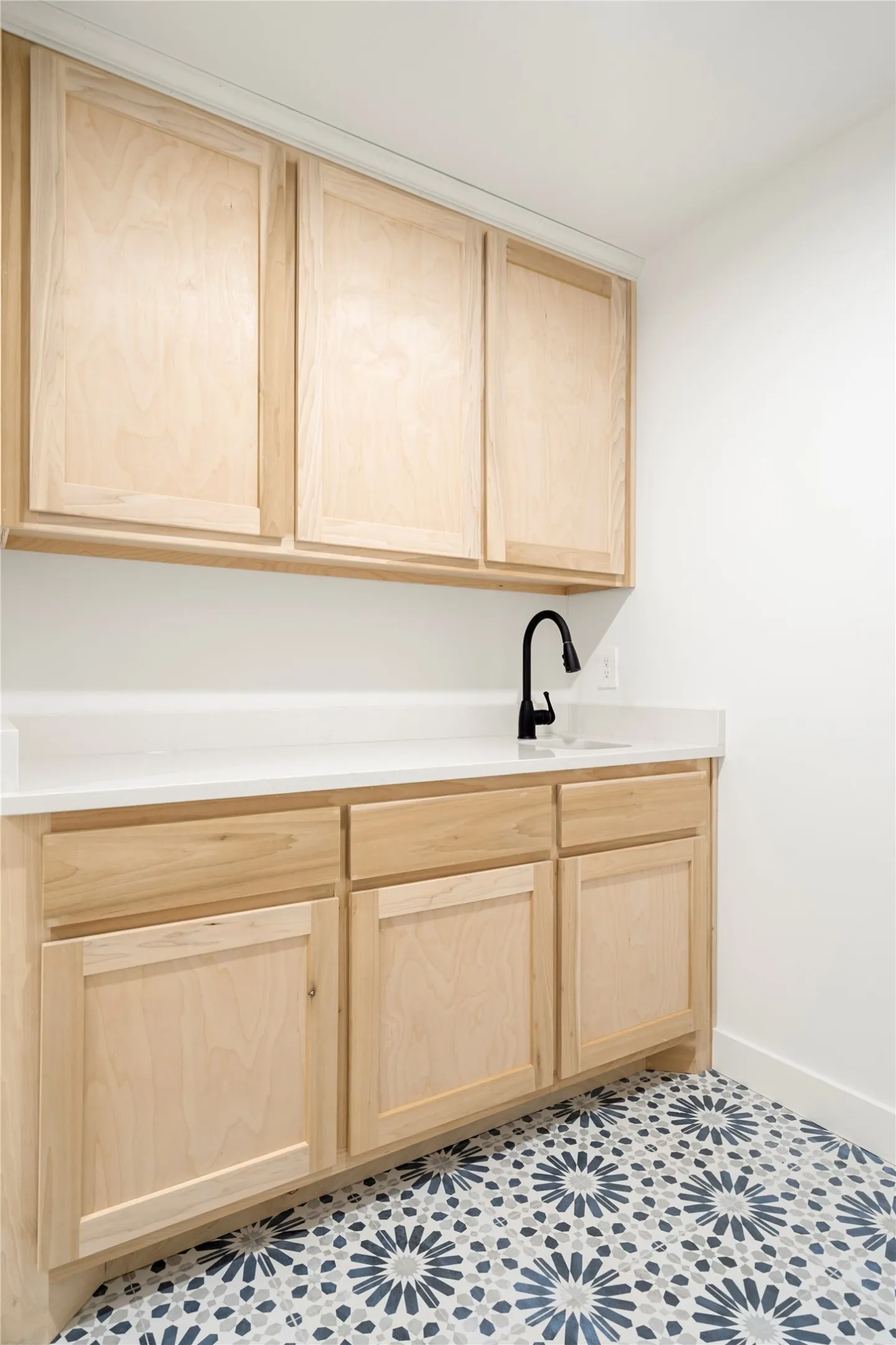 Light wood-finish cabinetry with white countertops, a black wall-mounted faucet, and a patterned tile floor