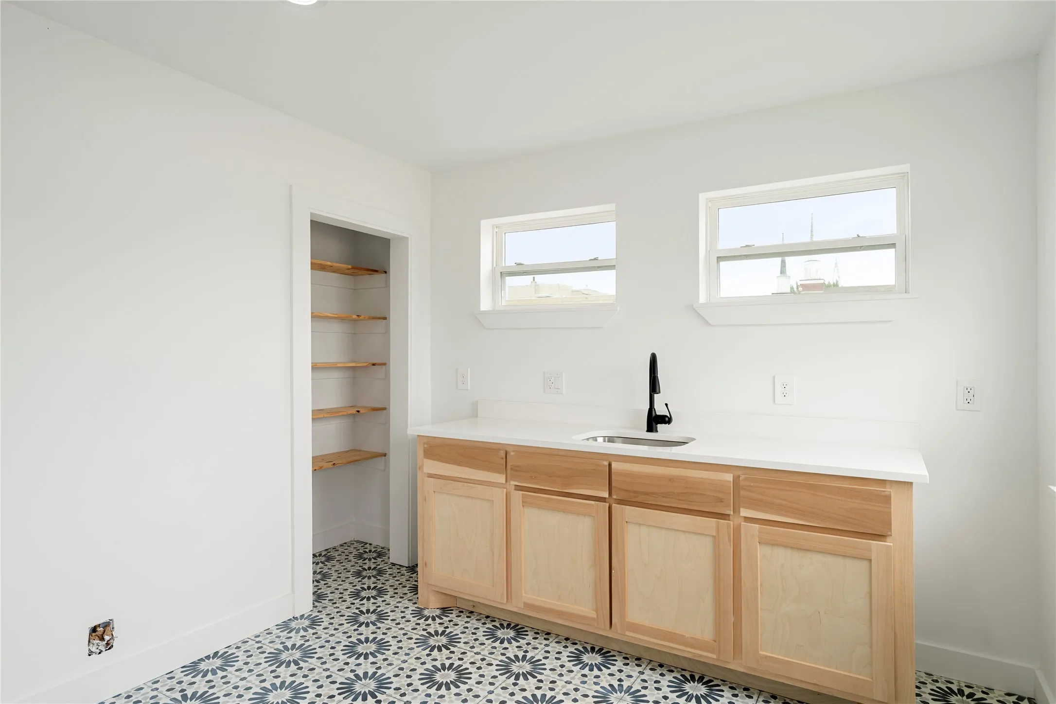Utility area featuring patterned tile flooring, light wood-finish cabinetry, a built-in sink with a matte black faucet, and two windows providing natural illumination