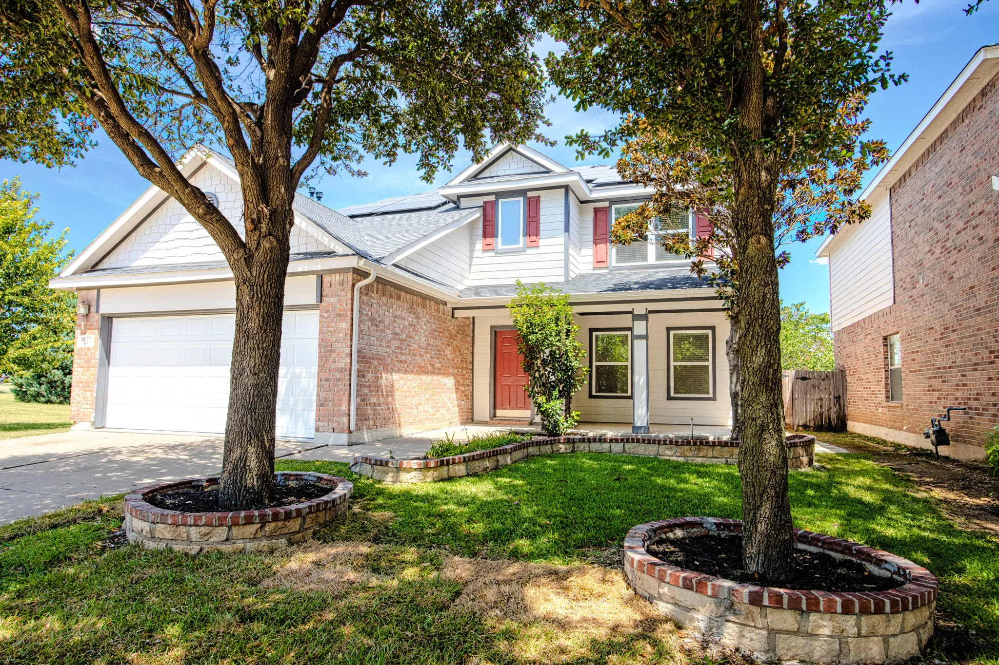 Two-story home featuring brick and siding exterior, a two-car garage, and mature trees with brick-edged planters