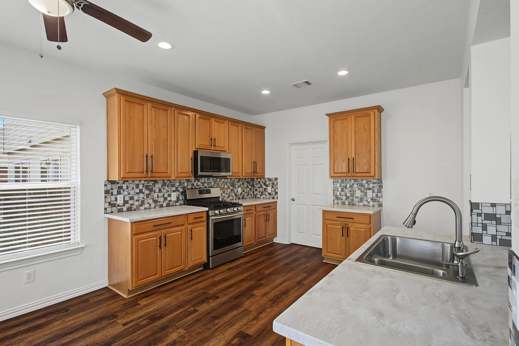 Warm wood-finish cabinetry, stainless steel appliances, patterned tile backsplash, wood-finish flooring, and a stainless steel sink with a gooseneck faucet
