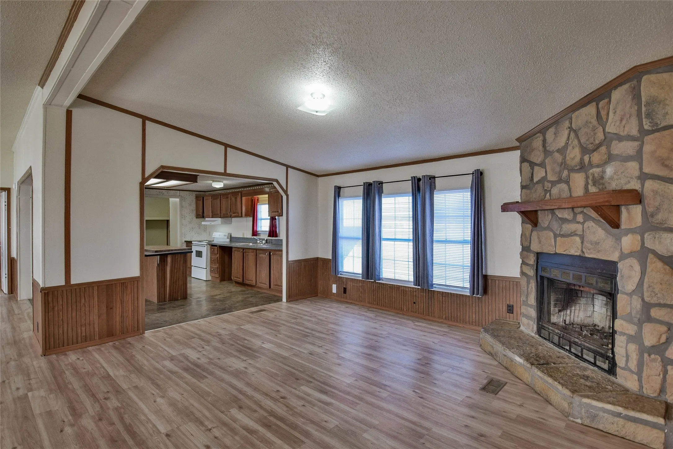 Wood-look vinyl flooring throughout, stone-clad fireplace with mantel, white kitchen appliances, and wood cabinetry