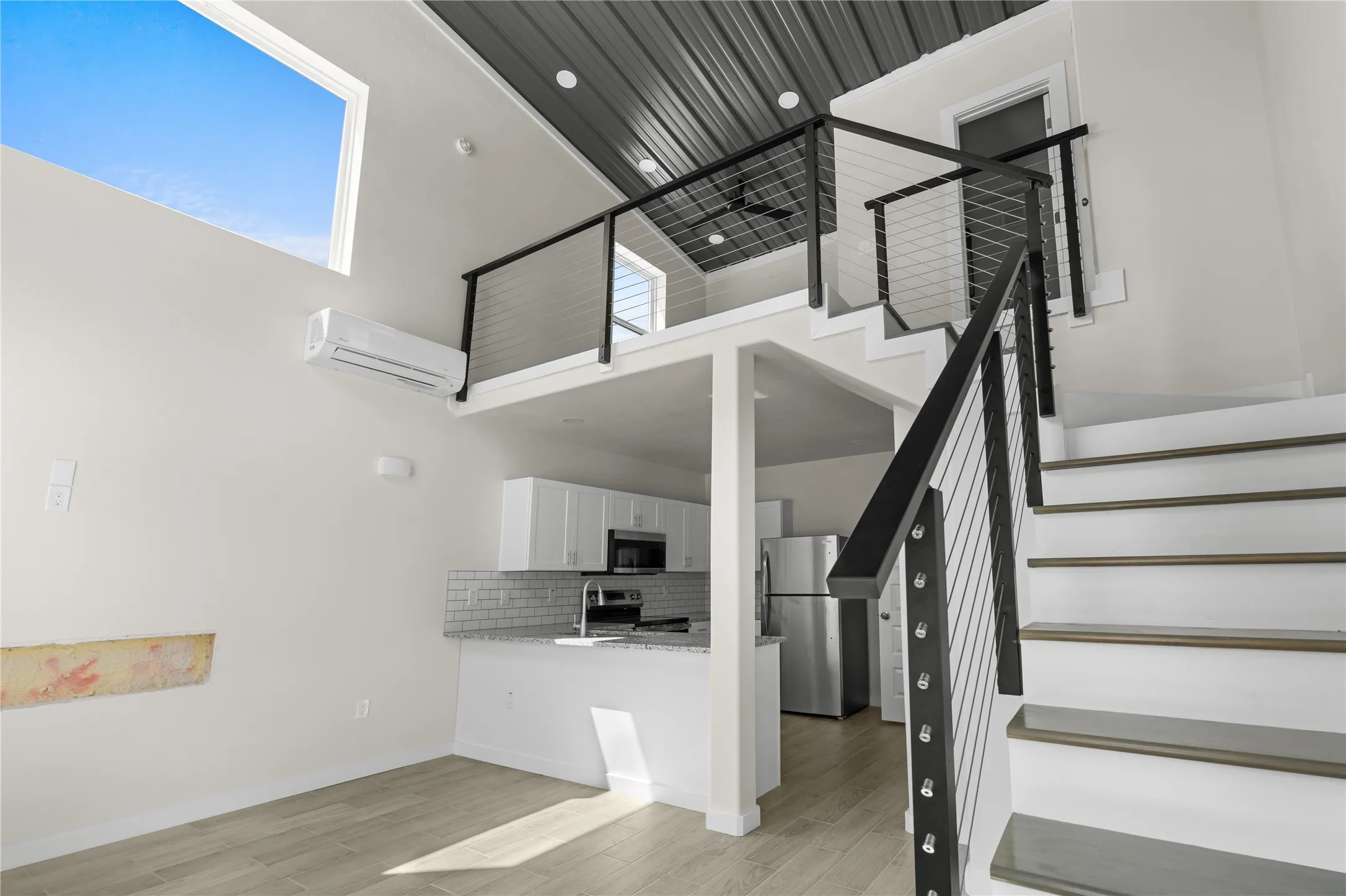 Soaring ceiling with a large transom window, modern cable railing on staircase and loft, stainless steel appliances, and white shaker cabinetry with subway tile backsplash