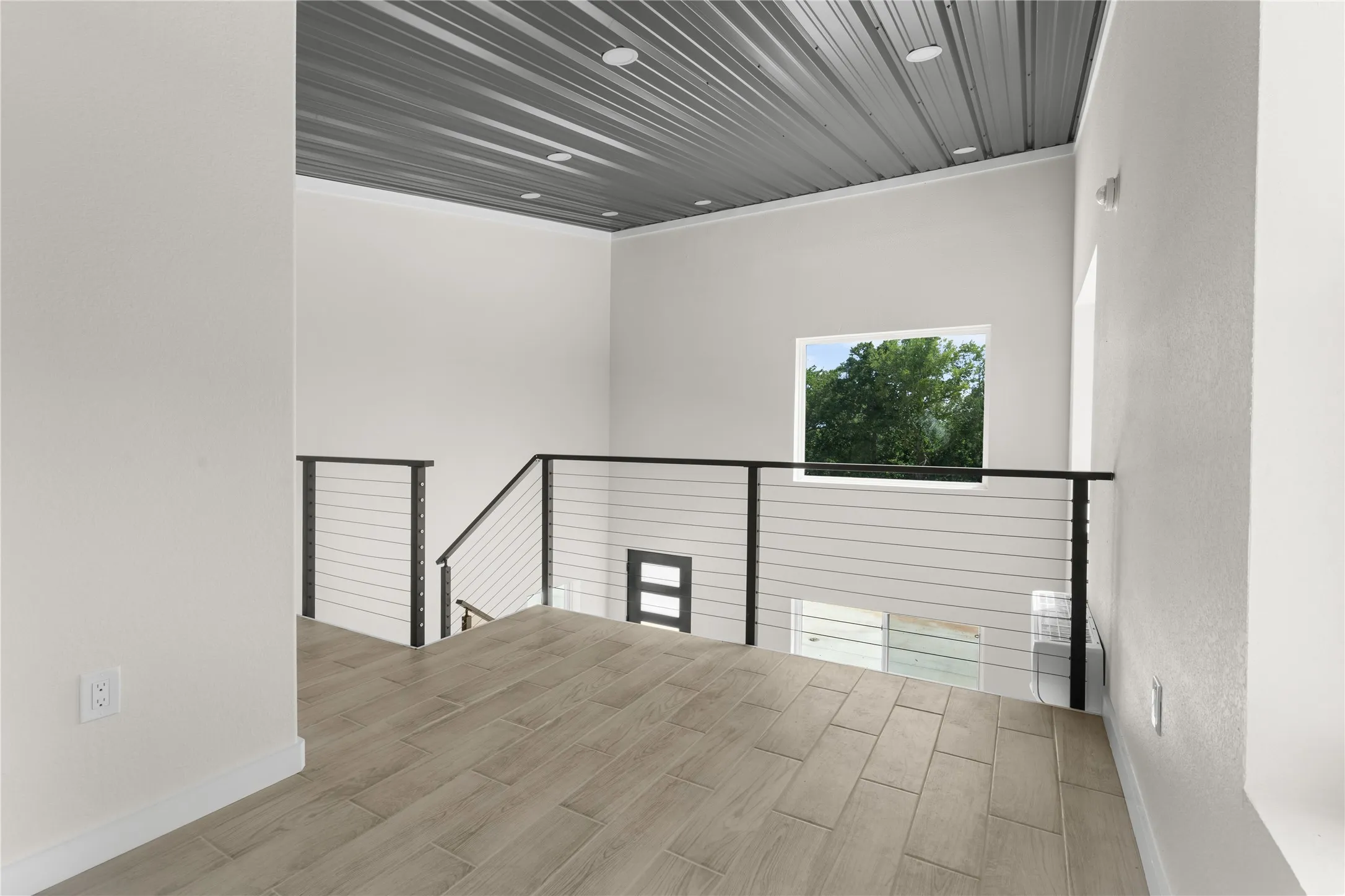 Modern cable railing system, wood-finish flooring, vaulted ceiling with recessed lighting, and a prominent window