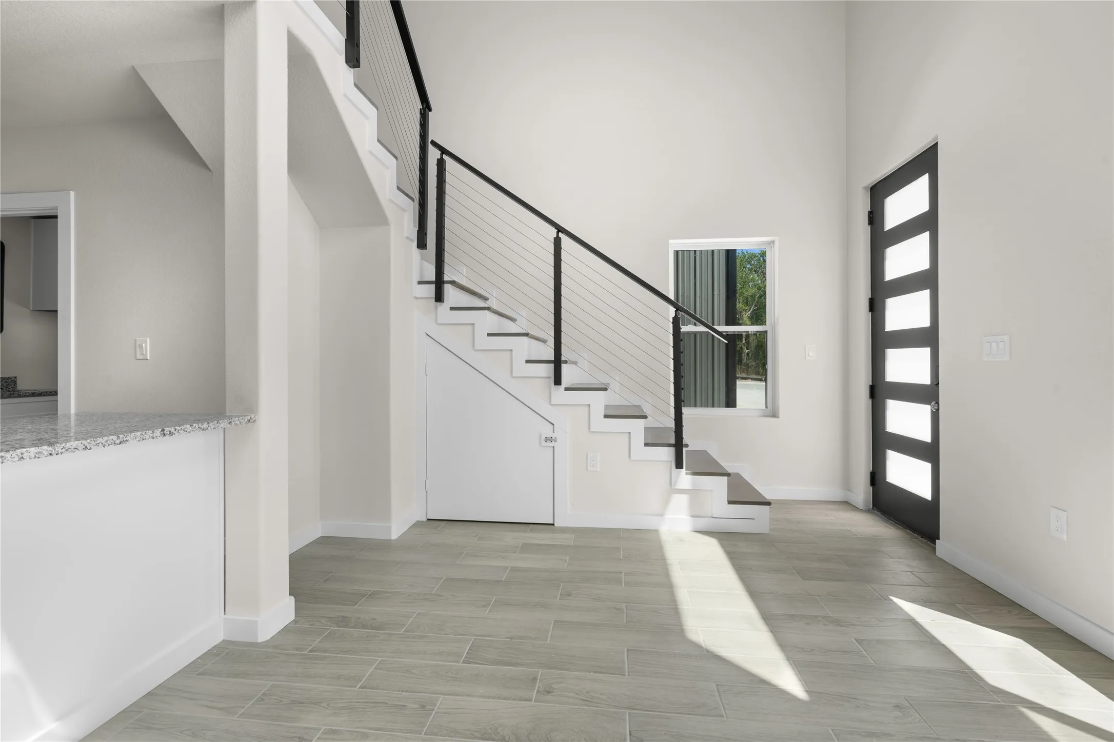 Vaulted ceilings, wood-finish flooring, staircase with metal railings, contemporary entry door with frosted panels, and a window providing natural illumination