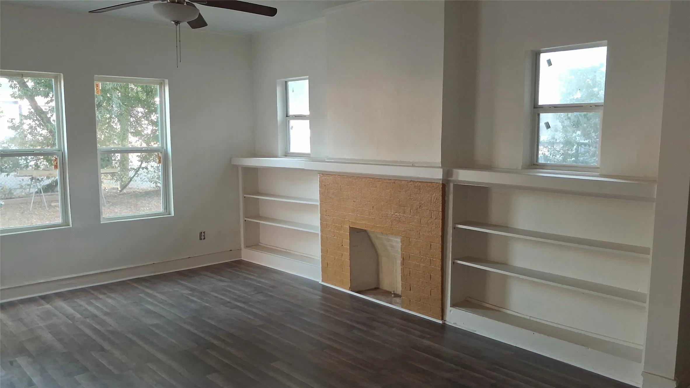 Wood-finish flooring, built-in shelving, textured fireplace surround, ceiling fan, and multiple windows