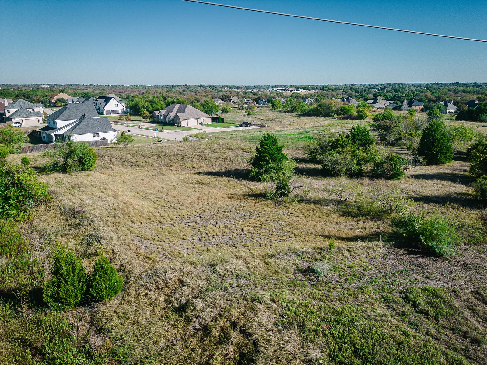 Expansive undeveloped land with scattered mature trees and varied ground cover, bordered by established residential properties