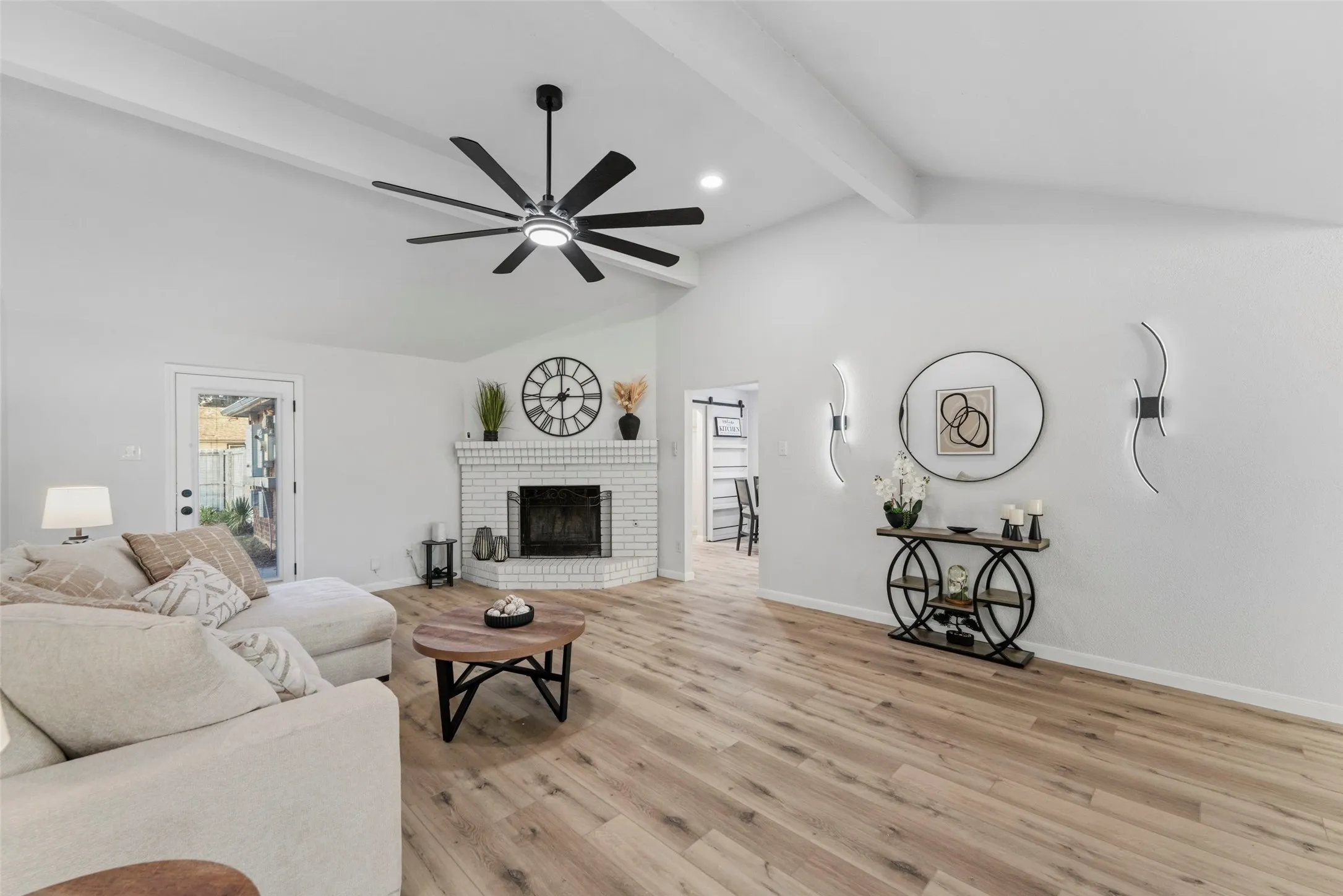 Vaulted ceiling with exposed beams, wood-finish flooring, white brick fireplace, modern ceiling fan, and decorative wall sconces