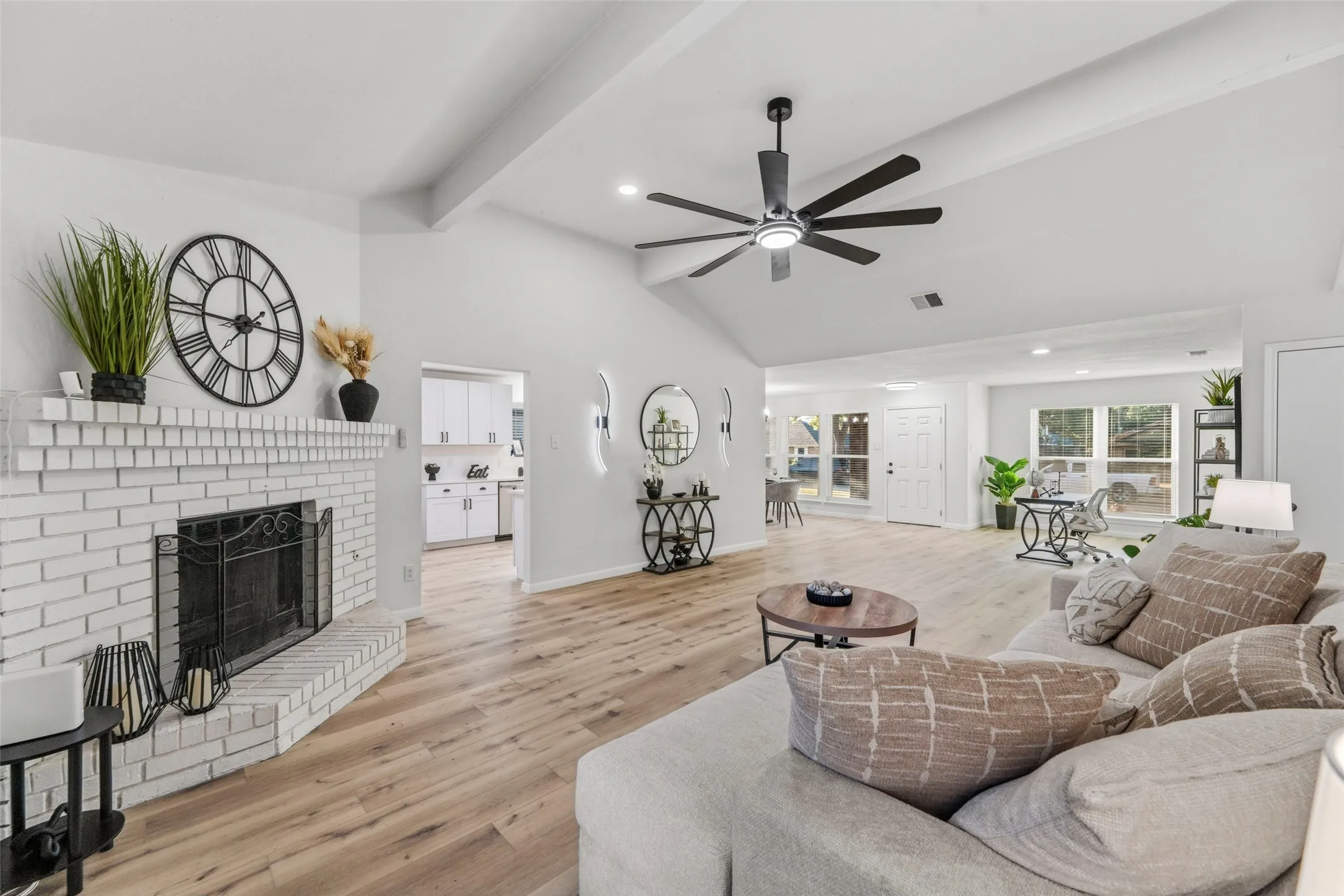 Vaulted ceiling with exposed beams, wood-finish flooring, white brick fireplace, modern ceiling fan, and recessed lighting