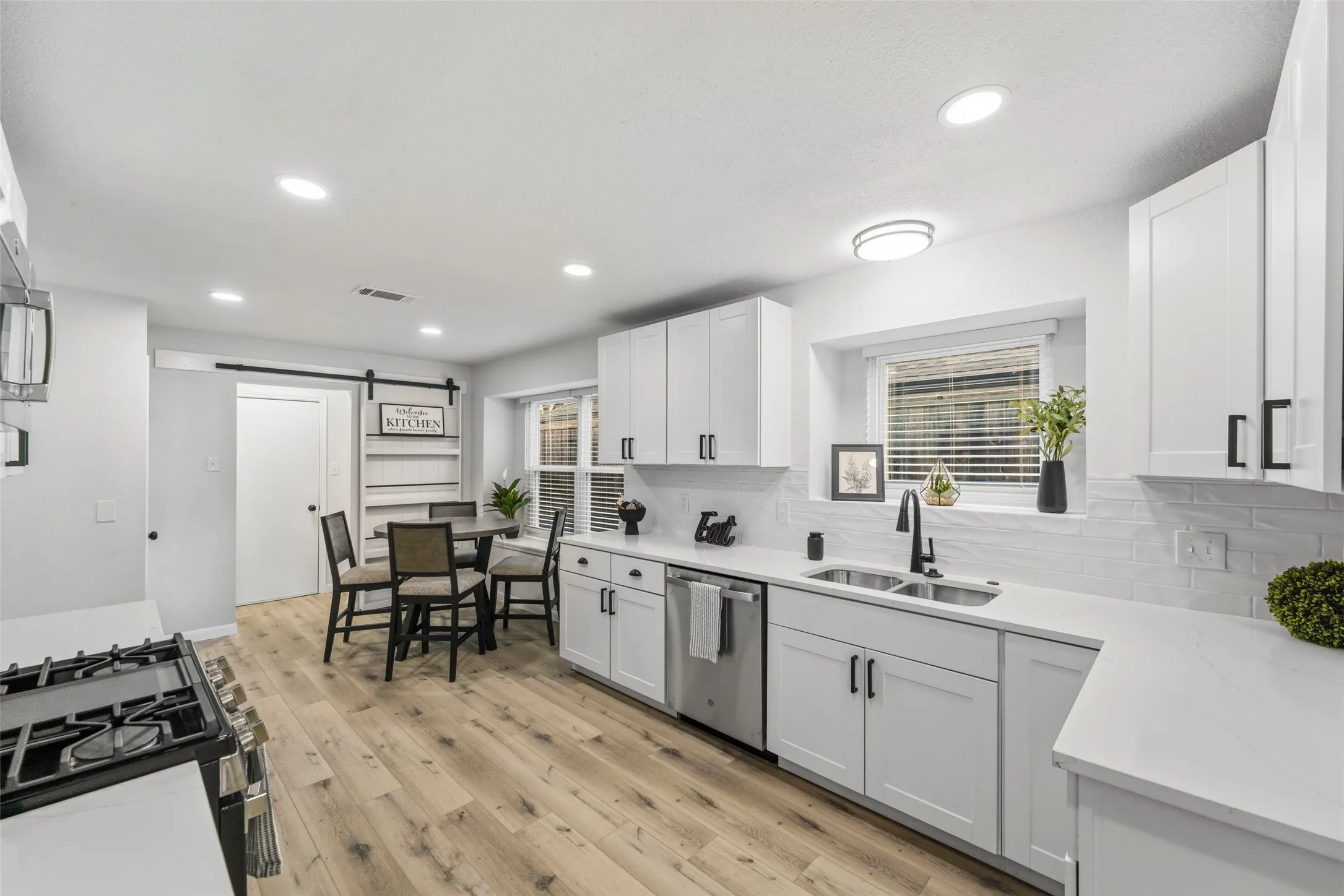 White cabinetry with matte black hardware, dual basin stainless steel sink, light wood-finish flooring, recessed lighting, and a barn door entry