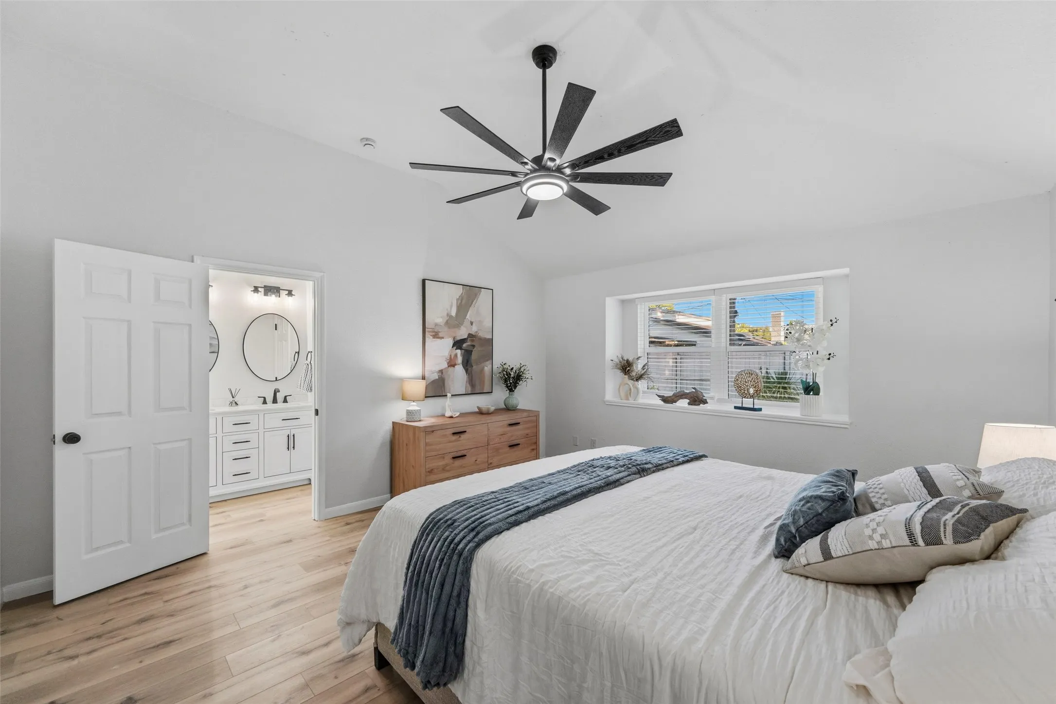 Vaulted ceiling with a contemporary ceiling fan, wood-finish flooring, a rectangular window with horizontal blinds, and an attached bath with a double vanity