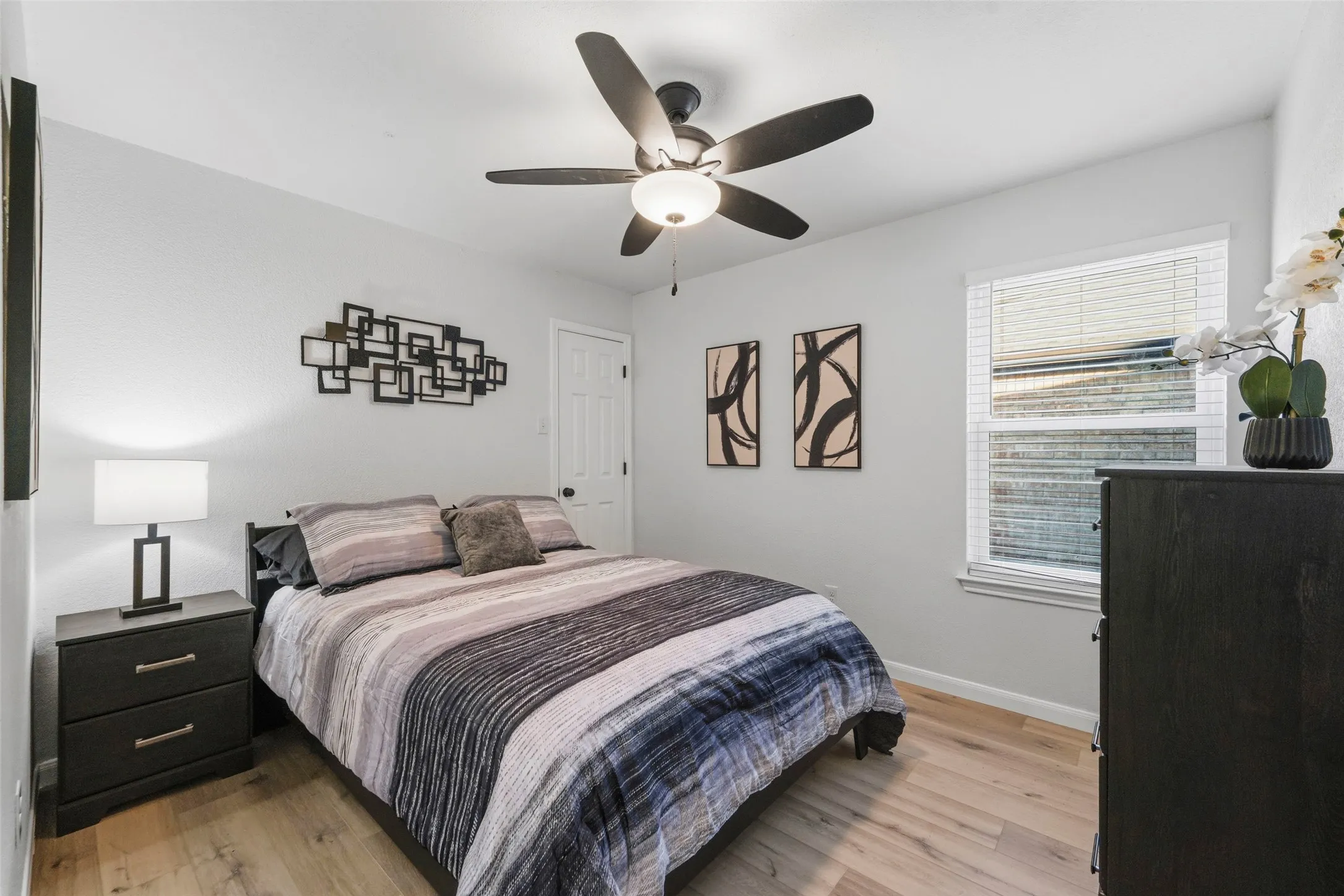 Light wood-finish flooring, white walls, ceiling fan with integrated lighting, and a white framed window with blinds
