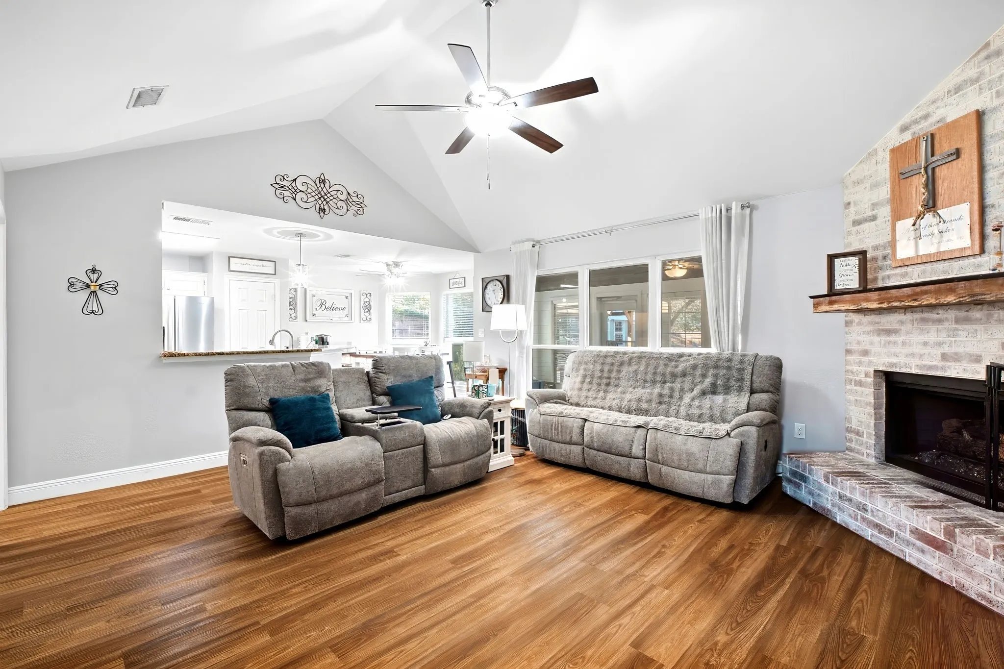 Vaulted ceiling with ceiling fan, wood-finish flooring, brick fireplace with wood mantel, pass-through window with countertop, and white painted walls