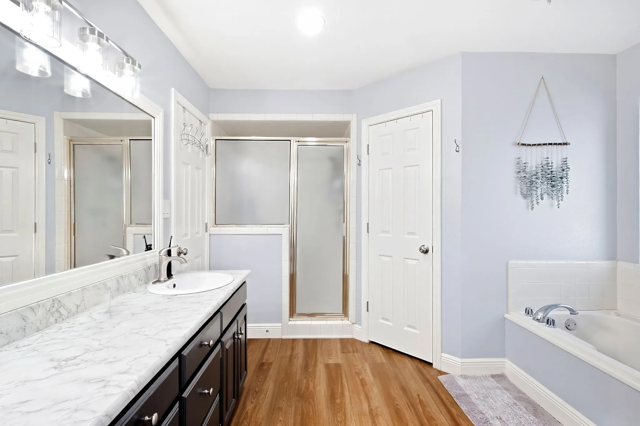 Bathroom featuring a single vanity with a white countertop, a built-in soaking tub, a shower enclosure with a frosted glass door, and wood-finish flooring