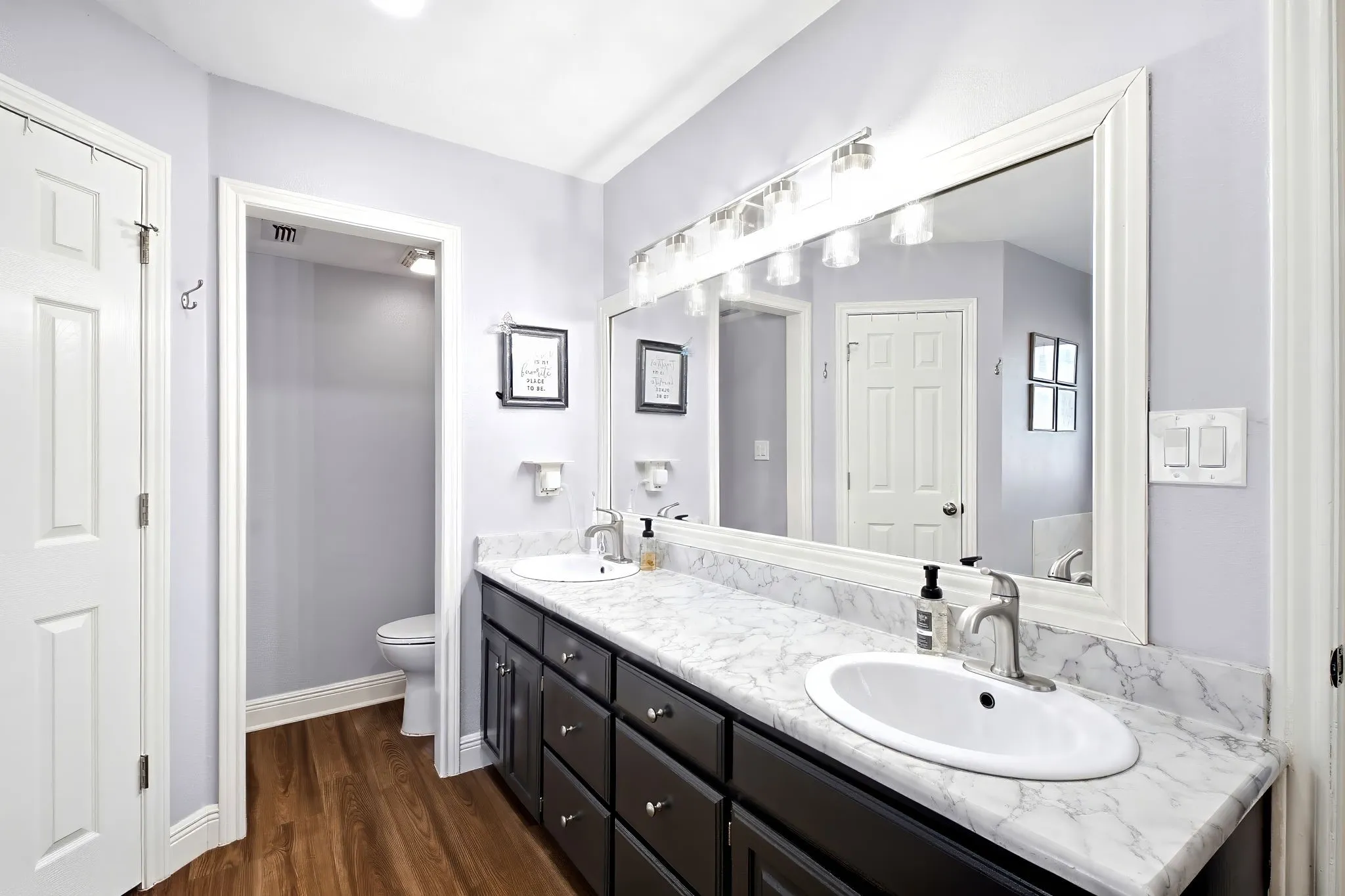Dual sink vanity with dark wood-finish cabinetry, white framed mirror, and wood-finish flooring