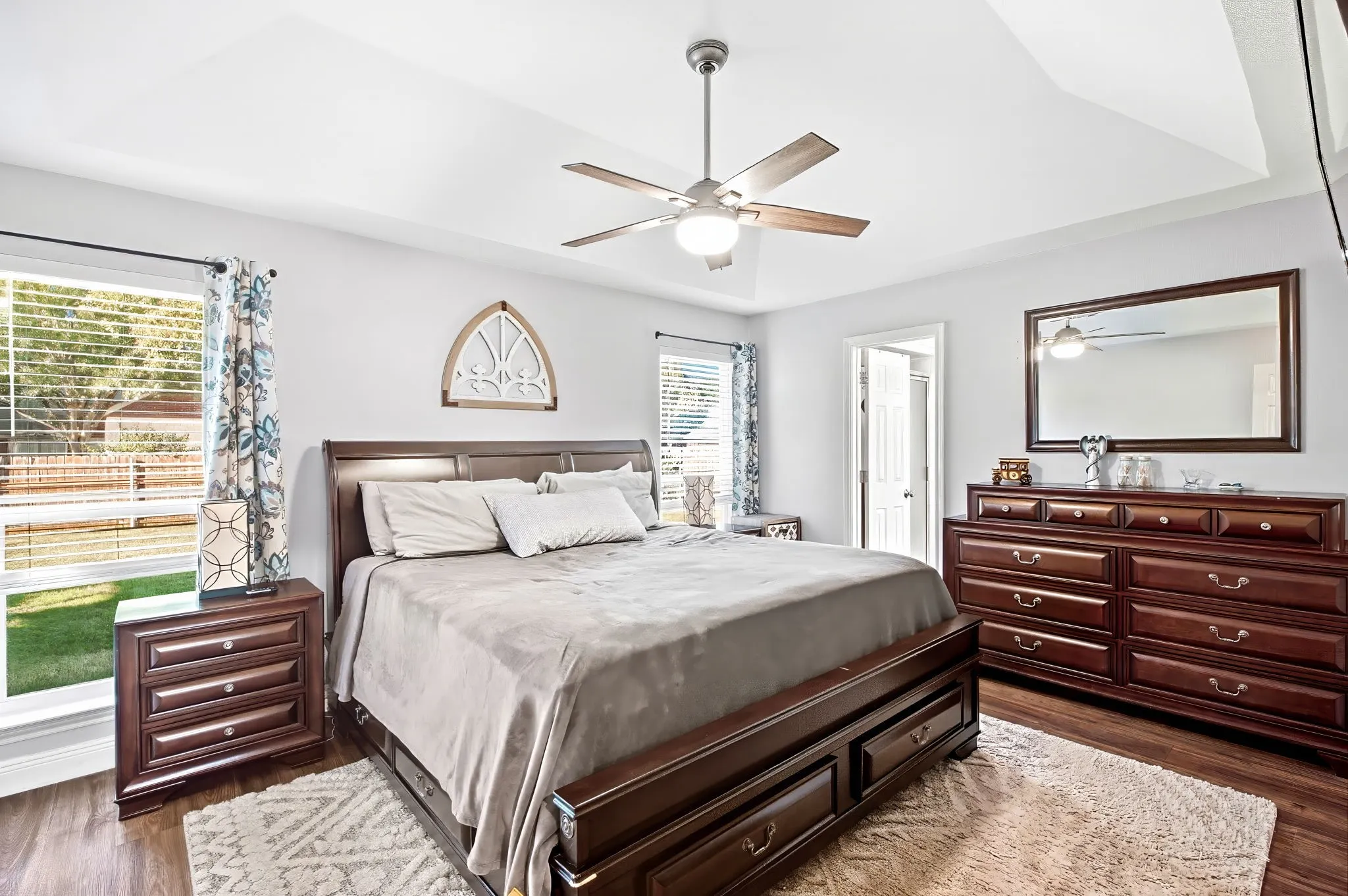 Tray ceiling, ceiling fan with light, wood-finish flooring, two large windows, and an interior doorway