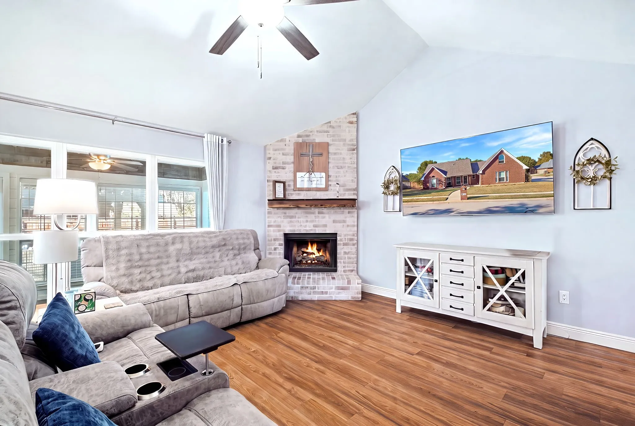 Vaulted ceiling with a ceiling fan, brick-clad fireplace with a wood mantle, wood-finish flooring, and large windows