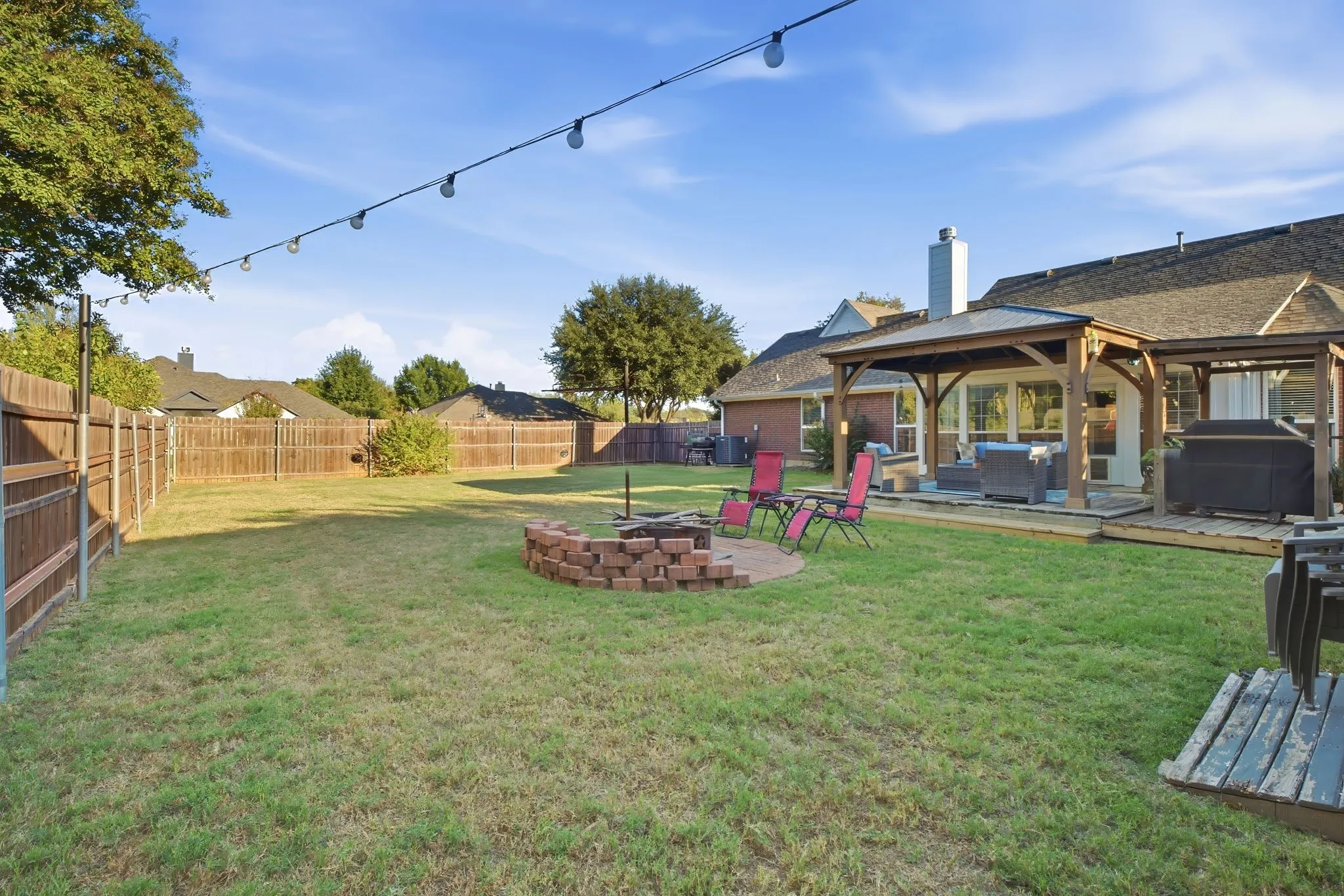 Expansive backyard featuring a manicured lawn, full perimeter fencing, a covered patio structure, and a brick-lined fire pit