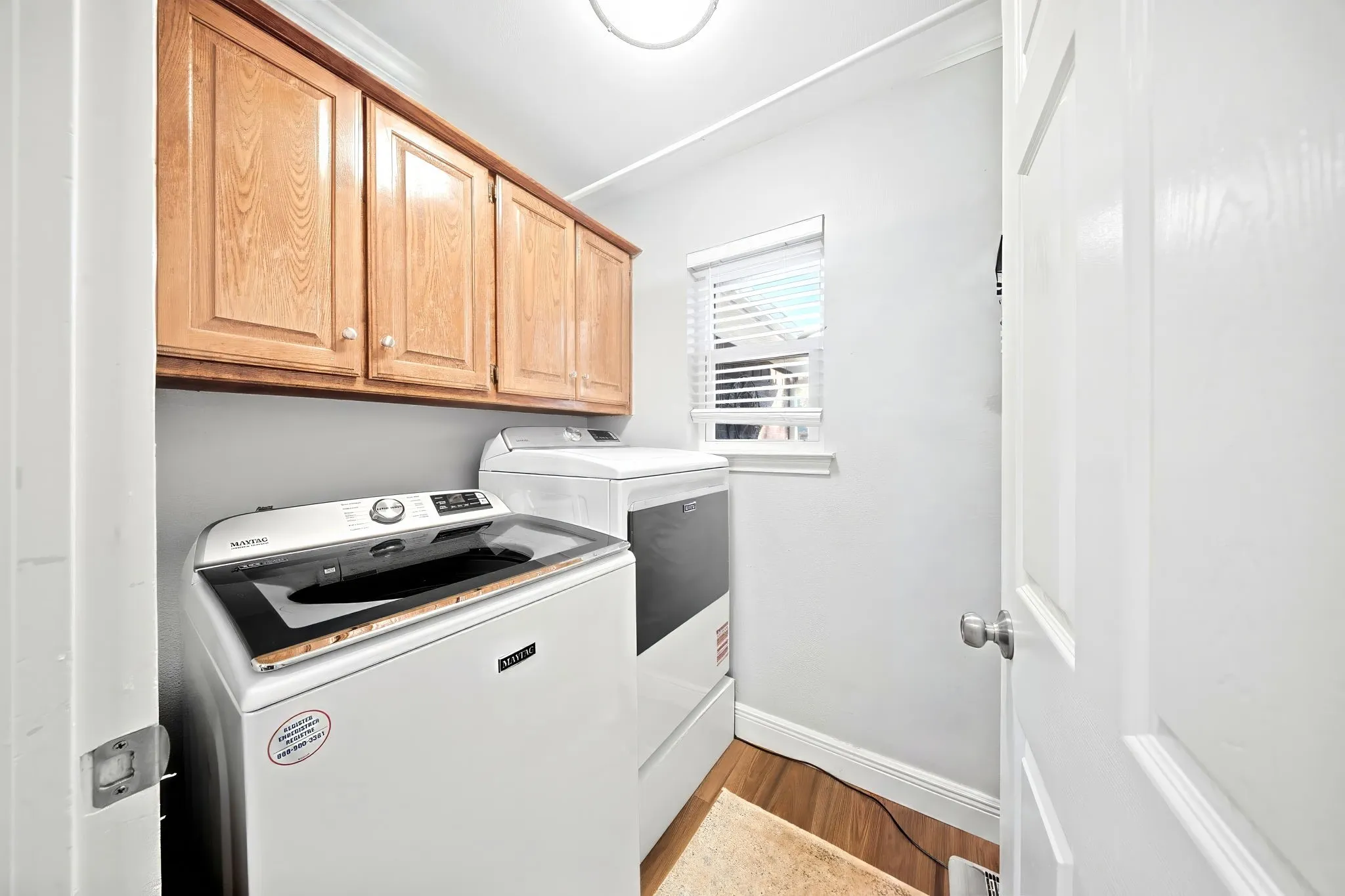 Laundry area featuring upper wood cabinetry, wood-finish flooring, a window with blinds, and white walls