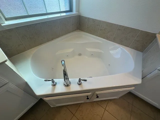 Corner jetted tub with chrome fixtures, surrounded by neutral tile backsplash and white cabinetry