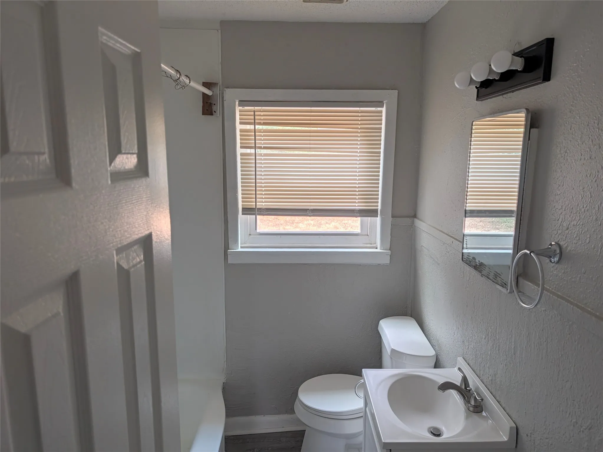 Window with horizontal blinds, vanity with integrated sink, wall-mounted mirror, three-light vanity fixture, and toilet