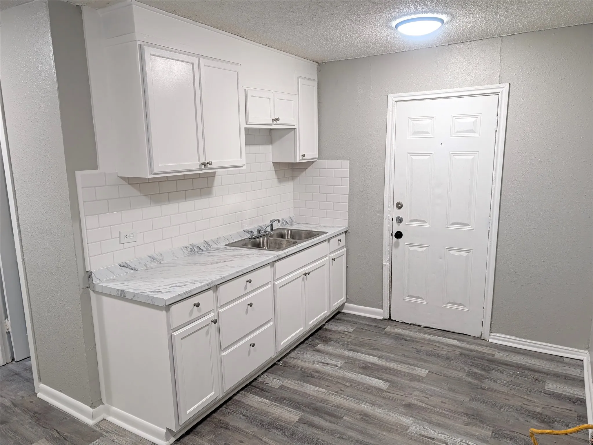 White cabinetry with brushed nickel hardware, white subway tile backsplash, light-toned countertop, double basin stainless steel sink, and wood-finish flooring