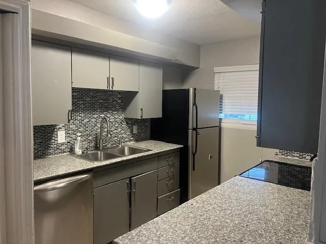 Kitchen featuring gray cabinetry, a mosaic tile backsplash, a double basin stainless steel sink, and stainless steel appliances