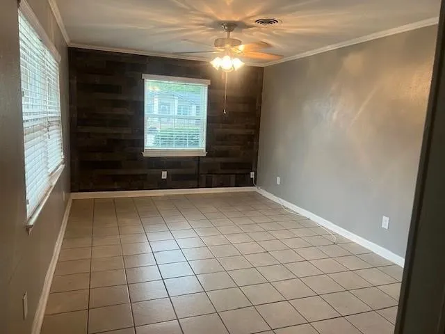 Tile flooring, accent wall with wood paneling, ceiling fan with integrated lighting, window with blinds, and crown molding