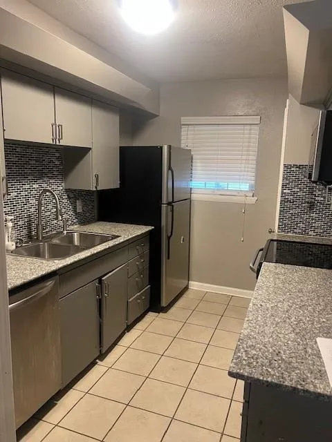 Kitchen featuring stainless steel appliances, dual basin sink, light-colored tile flooring, and speckled stone countertops